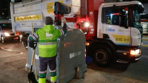Un trabajador está limpiando un contenedor mientras lo espera el camión de los residuos.