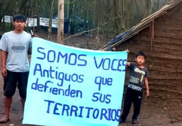 Hombre Mbya Guaraní sosteniendo un cartel que dice "Somos la voz de nuestros Ancestros" en un campamento de Misiones