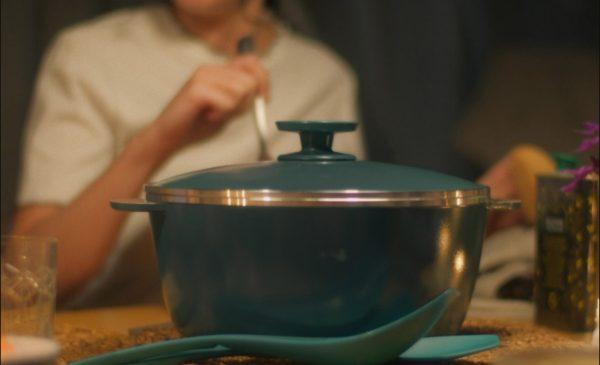 Una cacerola color verde, detrás una mano sosteniendo un utensilio de cocina