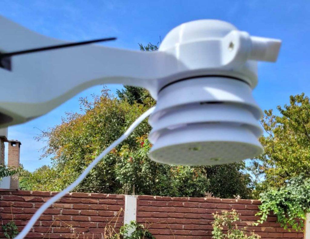 Plano en detalle de un sensor de calidad de aire color blanco, instalado en el exterior con vegetación, un muro de ladrillos y un cielo azul de fondo.