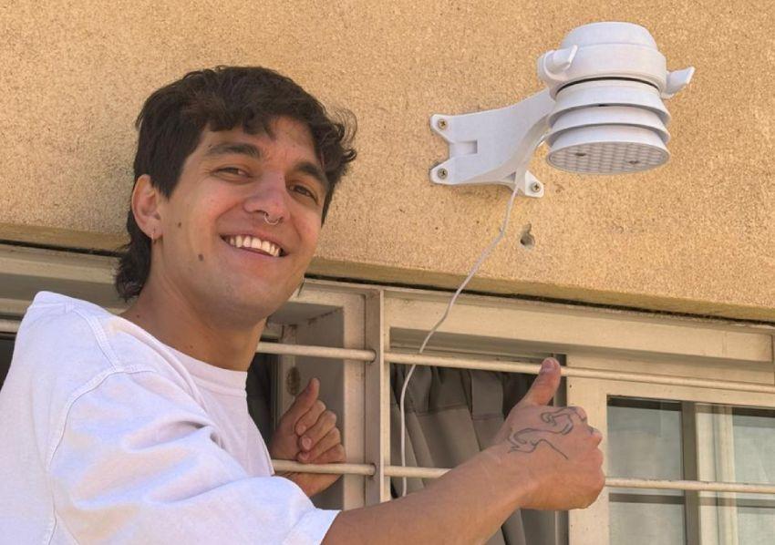 Un joven sonríe y hace un gesto de aprobación con el pulgar hacia arriba mientras muestra un sensor blanco de medición de aire montado en la pared de una vivienda.