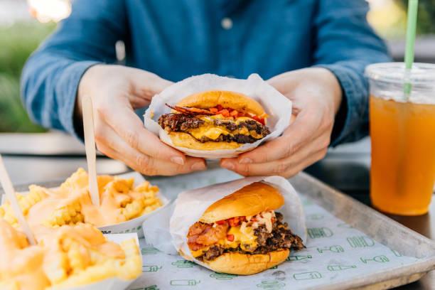 **Imagen 1 (Mesa con snacks y bebidas):** Vista aérea de una mesa repleta de alimentos ultraprocesados y botanas distribuidos en diversos cuencos, que incluyen papas fritas, palomitas de maíz, pretzels, cacahuates, nachos con salsas y aros de maíz, acompañados por vasos de refresco y cerveza con hielos y popotes. **Imagen 2 (Persona comiendo una hamburguesa):** Un hombre con lentes sostiene una hamburguesa con queso, jitomate y lechuga a punto de morderla. **Imagen 3 (Menú de comida rápida con hamburguesas y papas):** Una persona sostiene en sus manos una hamburguesa con queso y tocino envuelta en papel, servida en una bandeja metálica junto a otra hamburguesa similar, papas fritas bañado en salsa de queso y un vaso con bebida helada.