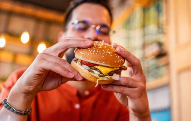 Un hombre con lentes sostiene una hamburguesa con queso, jitomate y lechuga a punto de morderla.