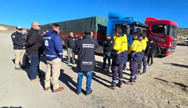 Grupo de funcionarios e inspectores en uniforme conversando en círculo junto a dos camiones de carga en un terreno descampado y soleado.