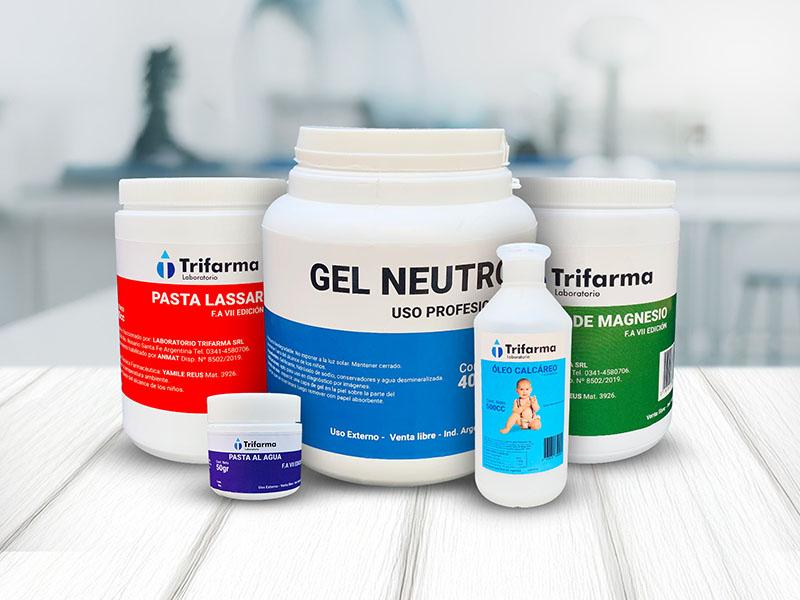 Conjunto de envases de productos farmacéuticos y cosméticos de la marca Trifarma sobre una superficie de madera clara, que incluye: un pote grande de "Gel Neutro", tarros de "Pasta Lassar", "Pasta al Agua" y "Hidróxido de Magnesio", y una botella de "Óleo Calcáreo" con la imagen de un bebé.