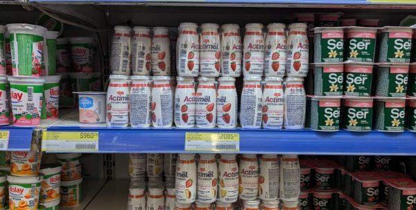 Estantes de supermercado repletos de lácteos, destacando filas de botellas blancas de Actimel sabor frutilla al centro, junto a yogures Ilolay y Ser con etiquetas de precio debajo.