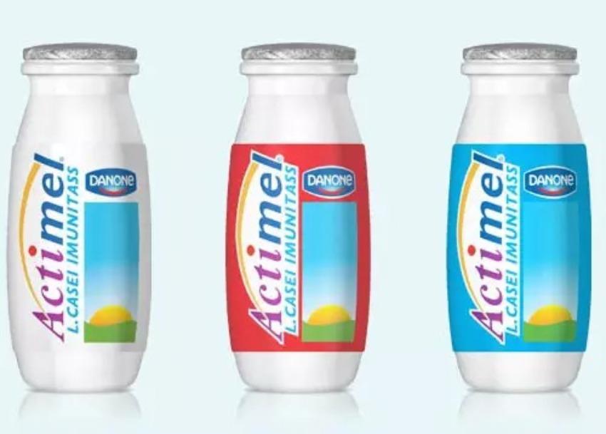 Tres pequeñas botellas de plástico blanco del lácteo Actimel de Danone ordenadas en fila sobre un fondo claro. Cada una tiene una etiqueta de color distinto alrededor del centro (gris claro a la izquierda, roja al centro y azul claro a la derecha), exhibiendo el logotipo vertical de Actimel, la marca Danone y la leyenda *"L. CASEI IMUNITASS"*.