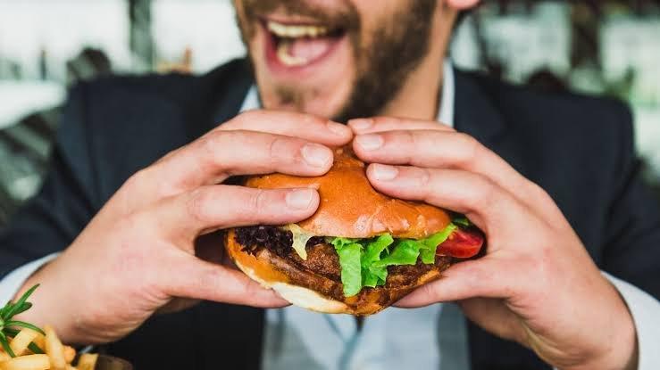 Dos manos agarran una hamburguesa. De fondo, la cara de un hombre con barba.