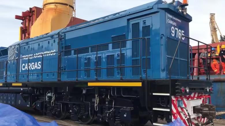 Parte de una locomotora azul con trabajadores en el margen derecho y otras máquinas ferroviarias de fondo