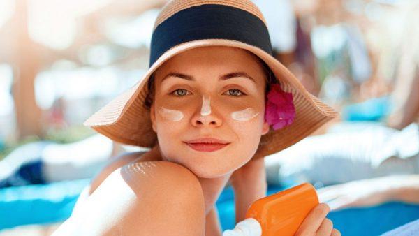 una chica tiene protector solar en la cara, un gorro de playa en la cabeza y el pote del protector en una de las manos