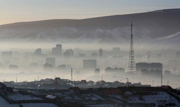 Panorama urbano de una ciudad parcialmente cubierta por una densa capa de niebla o smog, con montañas nevadas al fondo y una torre de telecomunicaciones en primer plano.