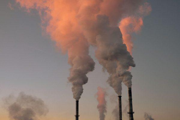 Texto alternativo (alt text) para la imagen: Tres chimeneas industriales emiten espesas columnas de humo denso hacia un cielo atardecido con tonos rojizos y anaranjados, ilustrando la contaminación atmosférica y las emisiones industriales.