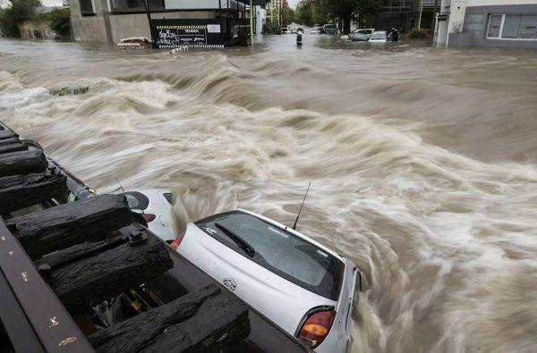 **Imagen (`el niño lluevia argentina.jpg`):** Fotografía de una inundación severa en una calle urbana, donde una corriente de agua rápida y con turbulencia arrastra o sumerge parcialmente varios automóviles. En primer plano se ven vías de tren o maderas estructurales a la izquierda y un auto compacto plateado casi cubierto por el agua turbia.