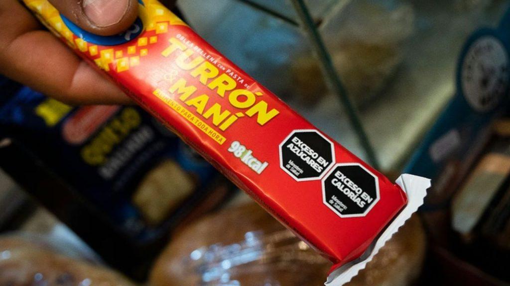 una mano sostiene un turrón de maní de Arcor que indica que tiene exceso de azúcares y en exceso en calorías