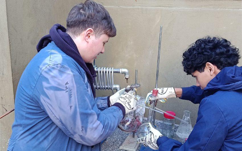Dos estudiantes con guantes de protección manipulan un equipo de laboratorio de vidrio con matraces y tubos de ensayo.