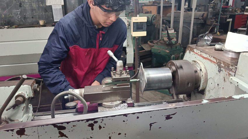 Un estudiante con gafas de protección opera un torno mecánico en un taller para moldear una pieza metálica
