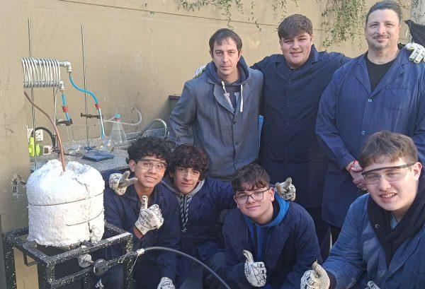 Un grupo de estudiantes y profesores posan sonrientes junto al prototipo de destilación de plástico en un patio al aire libre.