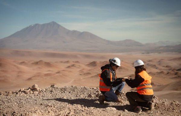 Dos trabajadores mineros, vestidos con chalecos de seguridad naranjas y cascos blancos, se arrodillan juntos en un peñasco rocoso para examinar un dispositivo o documentos. Al fondo se despliega un árido paisaje desértico con colinas arenosas y una montaña bajo un cielo despejado.