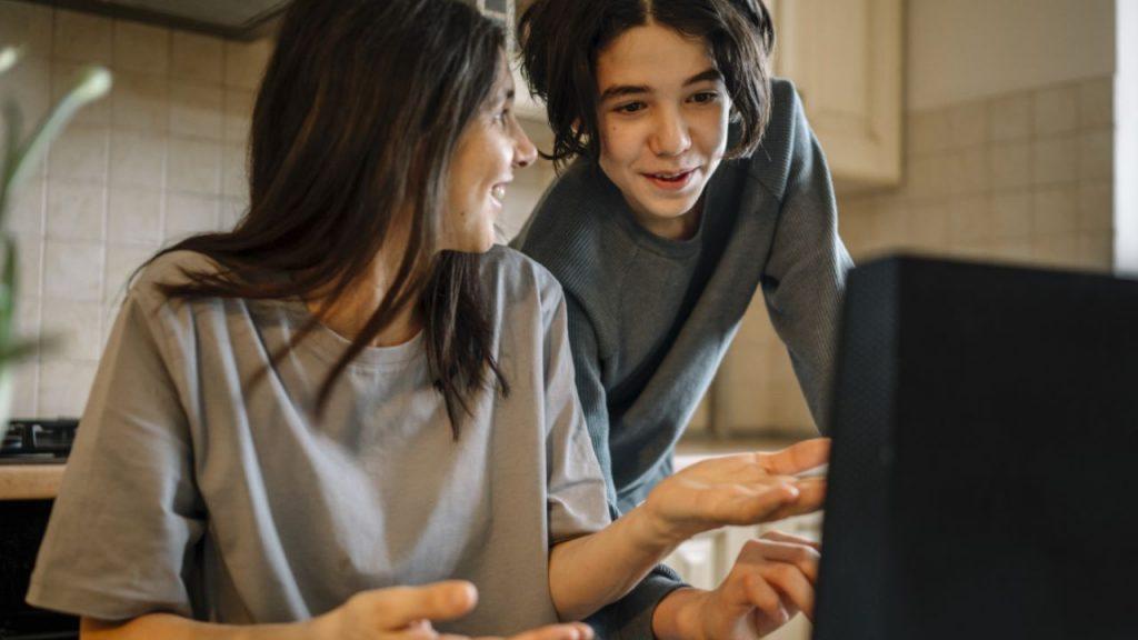 una madre se rie con su hijo mientras lo mira y él observa la pantalla de la computadora