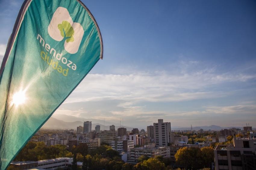 Bandera publicitaria con el logo de "Mendoza Ciudad" sobre una vista panorámica urbana al atardecer.