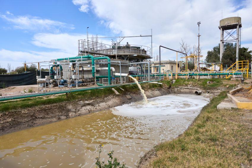 Vista general de una planta industrial de tratamiento de agua al aire libre, donde una tubería vierte un chorro de efluente turbio con espuma hacia un canal de tierra.
