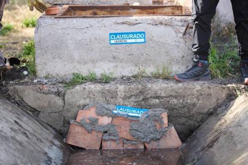 Vista en plano detalle de un canal de desagüe bloqueado con ladrillos y cemento, con dos fajas o pegatinas azules con la palabra «CLAUSURADO» pegadas tanto en la estructura superior como sobre el bloqueo.