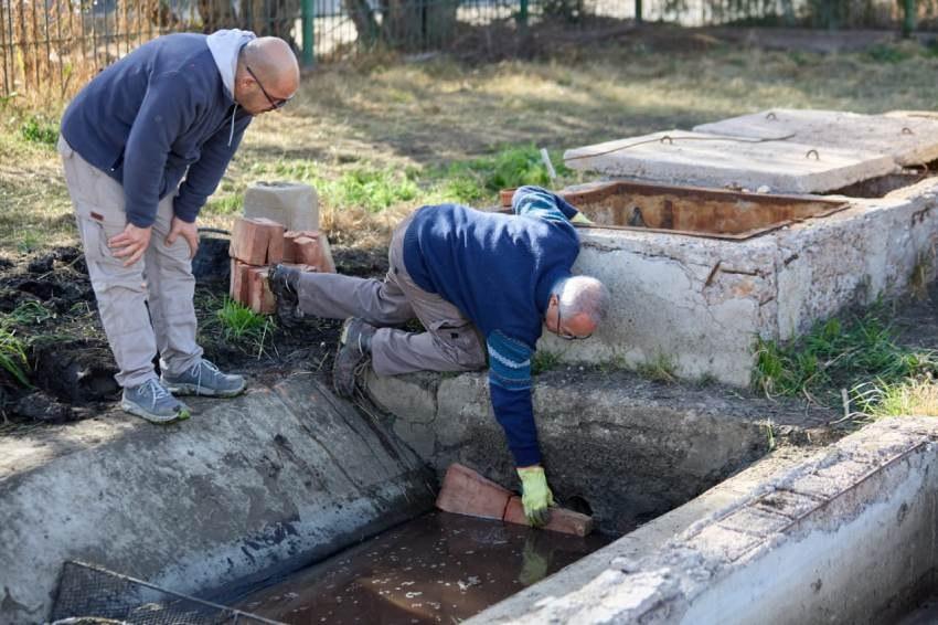 Dos operarios inspeccionan una infraestructura de desagüe en el exterior; uno de ellos está agachado sujetando un bloque dentro del canal de agua mientras el otro observa inclinado.