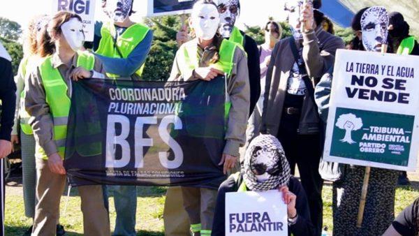 Grupo de manifestantes en un parque al aire libre, usando máscaras blancas y chalecos reflectantes, sostienen una bandera negra de la «Coordinadora Plurinacional BFS» junto a pancartas con consignas como «La tierra y el agua no se vende» y «Fuera Palantir».