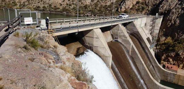 Vista en picado de una presa de hormigón entre cerros rocosos, con agua brotando por un vertedero y una camioneta blanca cruzando por la estructura superior.