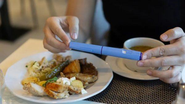Una persona sostiene con ambas manos un bolígrafo inyector de color azul sobre una mesa de comedor, teniendo enfrente un plato de comida servida y una taza.