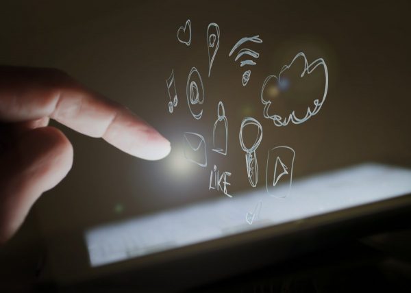Dedo de una persona apuntando hacia íconos ilustrados de redes sociales y tecnología digital que flotan sobre la pantalla de una tableta.