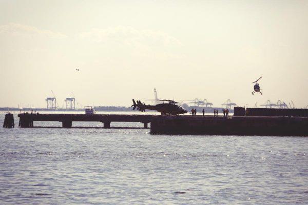 Un cuerpo de agua con leves olas ocupa la mitad inferior de la imagen. En el centro, un muelle de hormigón alberga un helicóptero posado junto a un pequeño grupo de personas en silueta. A la derecha, un segundo helicóptero más pequeño sobrevuela a baja altura cerca del borde del muelle.