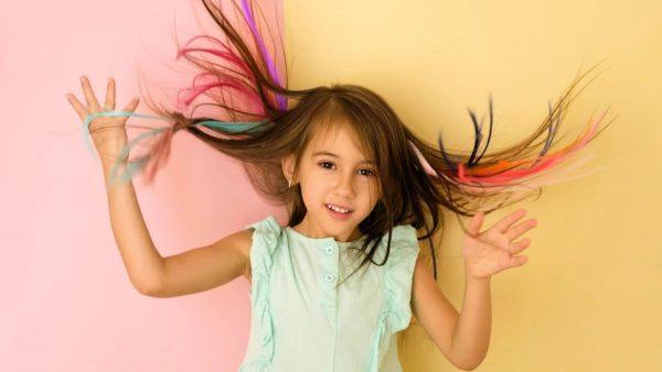 Una niña posa alegremente frente a un fondo dividido en dos colores pastel, rosa y amarillo. Lleva una blusa sin mangas de color azul claro y sostiene mechones de su cabello castaño claro hacia los lados, mostrando secciones teñidas de colores vibrantes como rosa, turquesa, azul y naranja.