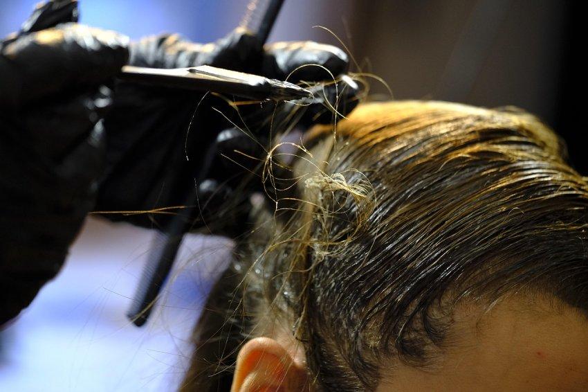 Un plano en primer plano de una persona en una peluquería mientras le aplican tinte o decolorante en el cabello con una brocha. La estilista lleva guantes negros de protección y peina el cabello húmedo y peinado hacia atrás, resaltando los tonos dorados y castaños bajo una iluminación enfocada.