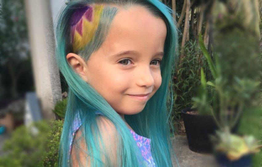 Una niña posa en un exterior sonriendo a la cámara, luciendo un peinado llamativo. Tiene el cabello largo de color turquesa o azul pastel con un rapado lateral en la zona de la sien, sobre el cual se aprecia un diseño teñido con patrones geométricos en tonos violeta y amarillo.