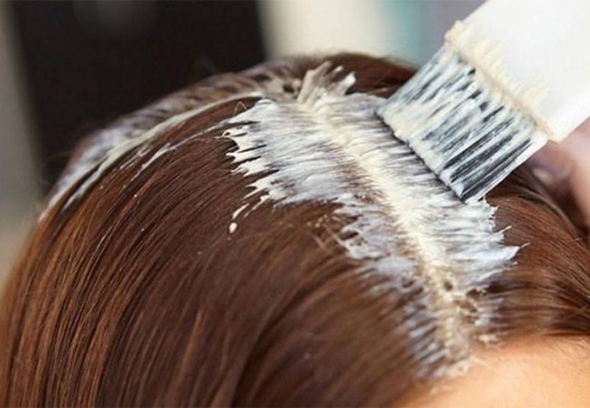 Un plano de cerca muestra la aplicación de un producto cremoso de color blanco o crema sobre la raya del cabello castaño de una persona. Una brocha de peluquería blanca distribuye el tinte o la mascarilla directamente a lo largo de la raíz.