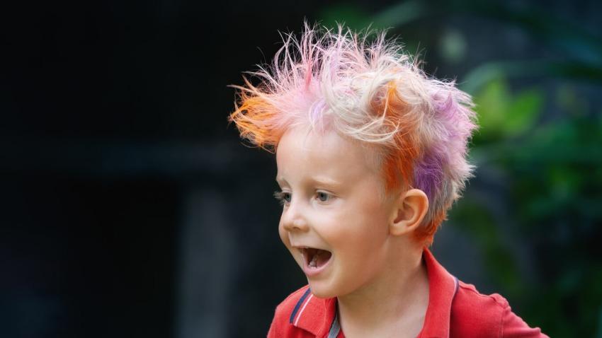 Un niño sonriente mira hacia un lado con expresión emocionada y la boca abierta. Lleva una chomba roja y luce un peinado corto y despeinado de puntas levantadas, teñido con una combinación vistosa de colores pastel y neón como rosa, naranja, morado y rubio claro.