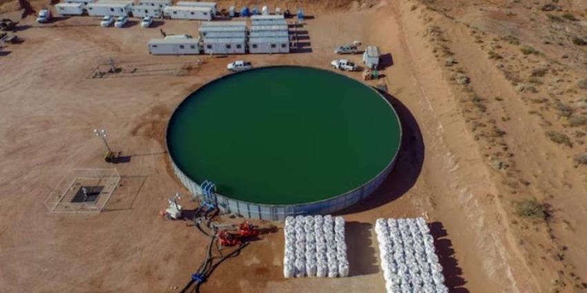 Vista aérea de un gran tanque circular de agua con líquido verdoso, rodeado de bolsas de insumos y campamento de tráileres en un yacimiento petrolero.