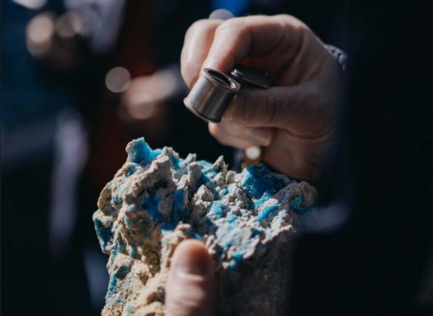 Una persona inspeccionando minuciosamente con una lupa de joyero un trozo de roca o mineral rugoso, el cual presenta incrustaciones y vetas de color azul brillante intenso.