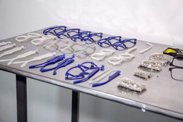 Marcos y patillas de anteojos de plástico azul, blanco y transparente ordenados sobre una mesa metálica de trabajo junto a piezas pequeñas.
