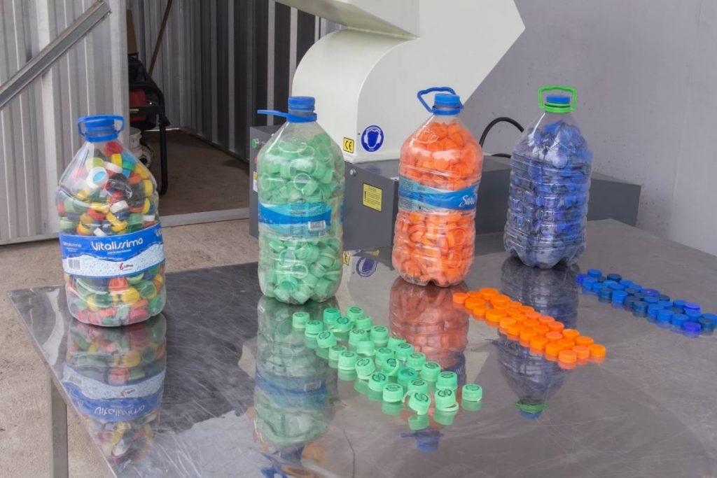 Botellones de plástico llenos de tapitas recicladas clasificadas por color y filas de tapitas alineadas sobre una mesa metálica frente a una máquina trituradora
