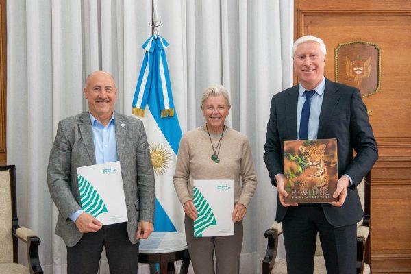 Tres personas posan de pie frente a la bandera argentina: a la izquierda, un hombre sostiene una carpeta de Parques Nacionales; en el centro, Kristine Tompkins; y a la derecha, un hombre trajeado muestra el libro "Rewilding en la Argentina"
