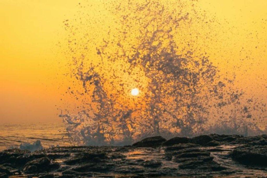 Fotografía del estallido de una ola del mar al romper contra rocas en la costa durante el amanecer o atardecer. La luz del sol tiñe el cielo y el agua con intensos tonos amarillos y dorados, con el disco solar brillando directamente a través de las gotas de agua proyectadas en el centro