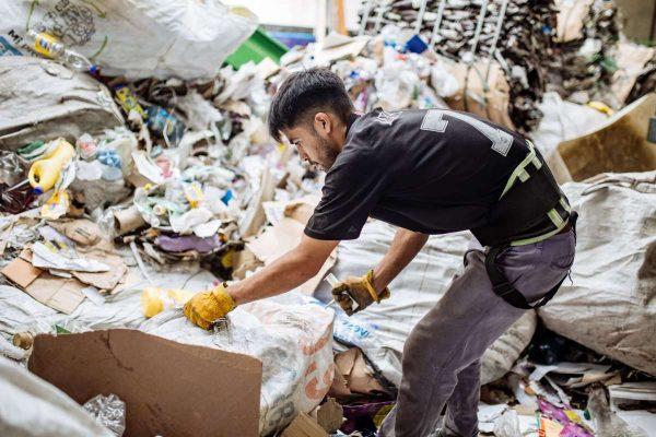 Texto alternativo: Joven con guantes de trabajo manipulando y clasificando materiales reciclables en una planta de separación de residuos.