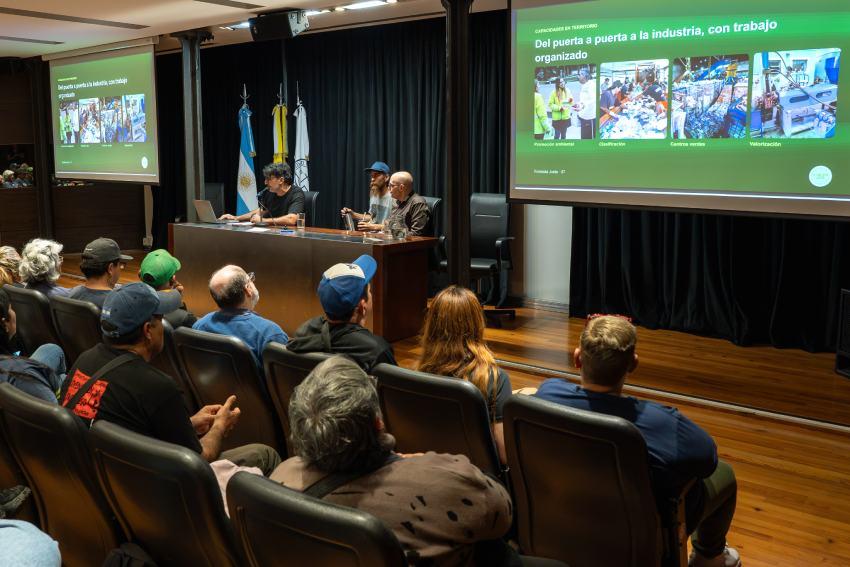 Asistentes en un auditorio de la UCA observando a los tres disertantes en el escenario y una pantalla que proyecta la diapositiva "Del puerta a puerta a la industria"