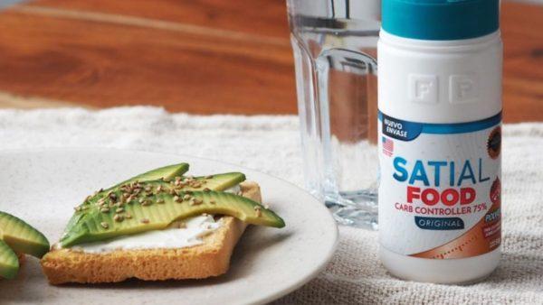 Envase de Satial Food junto a una tostada con queso untable, palta y semillas sobre un plato blanco, con un vaso de agua al fondo