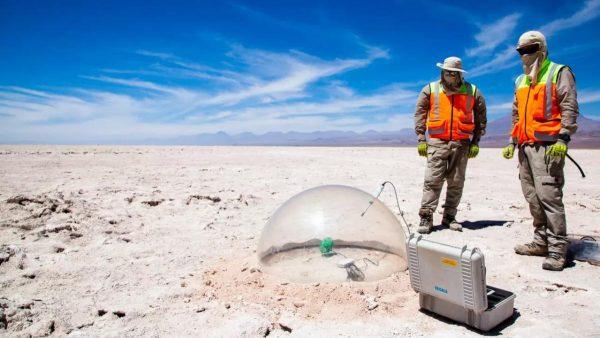 Dos trabajadores equipados con chalecos reflectantes y protección solar observan un domo transparente de medición instalado sobre el terreno árido de un salar, junto a un maletín de equipamiento técnico bajo un cielo despejado.