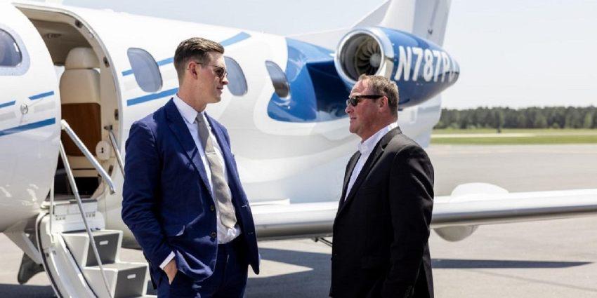 Dos hombres elegantes en traje y anteojos de sol conversan en la pista de aterrizaje junto a la escalera de entrada de un avión privado blanco y azul.
