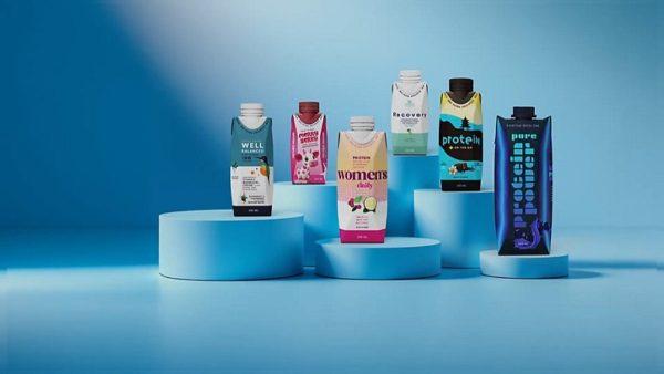 Varias botellas y envases de cartón de bebidas de proteínas de diferentes marcas y diseños coloridos dispuestos sobre soportes cilíndricos escalonados, con un fondo monocromático en tonos azules.