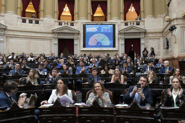 Fotografía desde el plano del recinto de la Cámara de Diputados. En primer plano y en las bancas se observa a varios diputados aplaudiendo y tomando fotografías. Al fondo, sobre la pared de arquitectura clásica con cortinados rojos, una gran pantalla azul despliega los resultados de la votación del proyecto de "Ley de Inviolabilidad de la Propiedad Privada": 124 votos afirmativos, 119 negativos y 6 abstenciones.